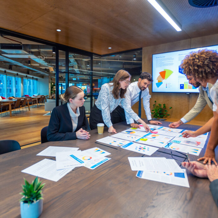 Paperwork and group of peoples hands on a board room table at a business presentation or seminar. The documents have financial or marketing figures, graphs and charts on them. People are pointing at the data. There is a T.V. with financial data in the background