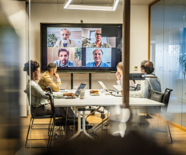 Business people attending a hybrid meeting with some people in a conference room and others attending via video conferencing on a large screen.